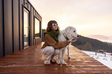 Woman hugs her dog while resting on terrace of tiny house in the mountains on winter time. Concept...