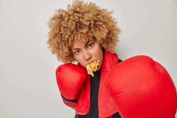 Fitness woman has mouth full of french fries stands in defensive pose exercises punches wears red...
