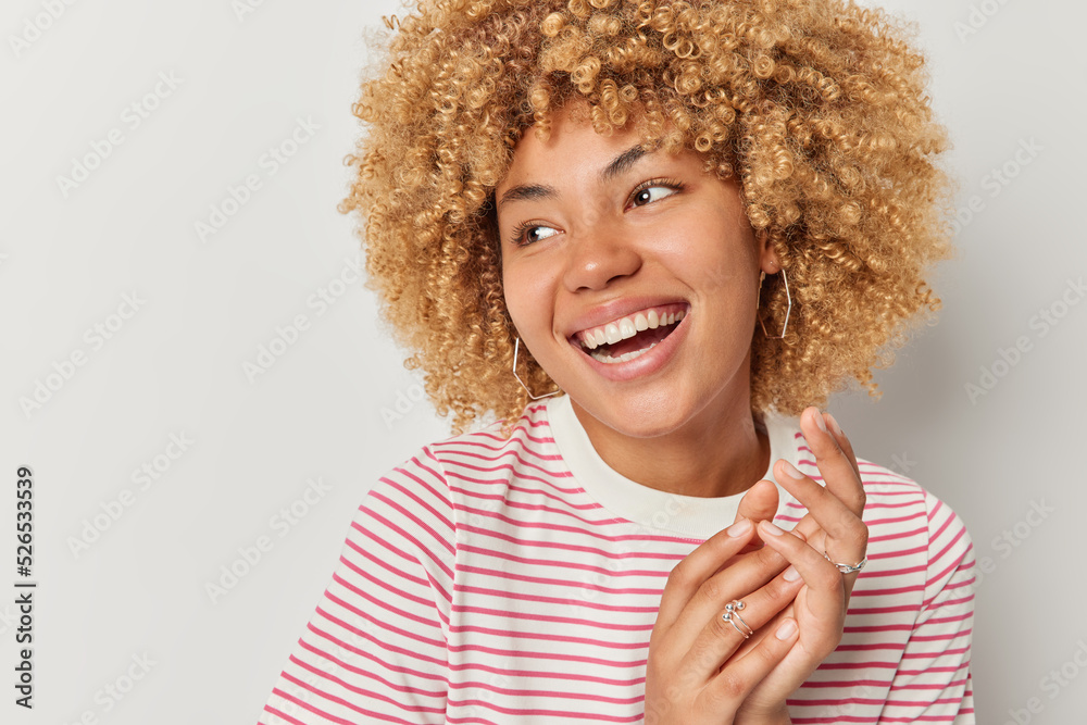 Wall mural Cheerful woman with curly hair rubs palms smiles toothily being in good mood dressed in casual striped t shirt concentrated aside isolated over grey studio wall with copy space aside. Happiness - Wall murals