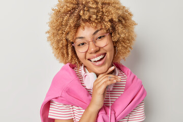 Beautiful curly haired woman smiles broadly keeps hand under chin focused forward expresses positive emotions wears big spectacles striped t shirt pink pullover tied over shoulders uses headphones