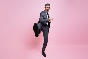 Furious mature man in formalwear throwing leg kick while standing against pink background