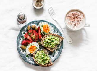 Delicious balanced breakfast - avocado toast, boiled egg, fresh tomatoes and cappuccino on a light background, top view