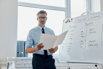 Confident English teacher holding papers while standing near the whiteboard at classroom