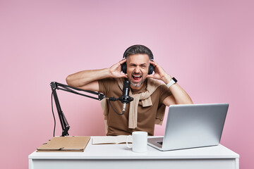 Excited man in headphones using microphone while recording podcast against pink background