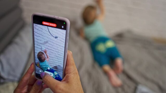 Mommy is taking video of her son on the smartphone. Little baby boy crawling by the bed exploring the white brick wall. Blurred backdrop.