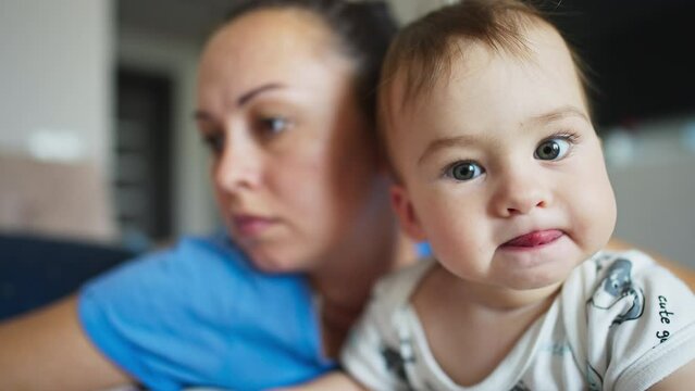 Mother and her baby boy beside. Little cite kid looks into camera with interest showing his tongue. Close up. Blurred backdrop.