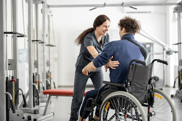 Fototapeta na wymiar Rehabilitation specialist helps a guy stand out of a wheelchair at rehabilitation center. Concept of physical therapy and support for people with disabilities