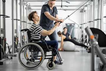 Rehabilitation specialist helps a guy in a wheelchair to do exercise on decompression simulator for recovery from injury. Concept of physical therapy for people with disabilities - 526539347