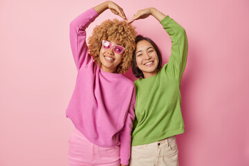 Obraz na płótnie Canvas Two beautiful women raise arms and shape heart smile gladfully dressed in casual pullovers and trousers have fun express positive emotions isolated over pink background. Friendship and relationships
