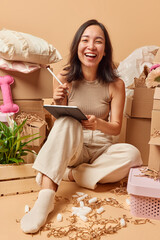 Vertical shot of cheerful young Asian woman poses in messy room relocates to new apartment uses touchpad and stylus thinks about design of house laughs happily poses on floor around carton boxes