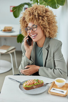 Elegant cheerful female in formal wear has positive phone conversation discusses good news poses at white table eats tasty sandwich and drinks tea makes call to best friend poses against cozy interior