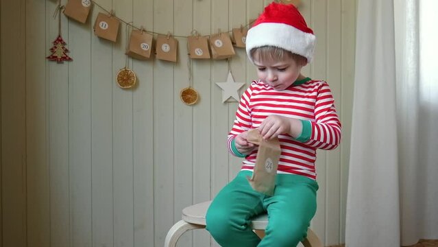 Boy in pajamas, Santa cap eating sweet candy on paper advent calendar with presents background. Little happy child with gift of handmade Christmas calendar on wall. Celebrating at home Xmas tradition.