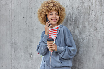 Fototapeta premium Young glad woman with curly hair makes phone call enjoys pleasant mobile conversation holds takeaway coffee focused aside dressed in casual hoodie poses over grey concrete wall. Distant communication