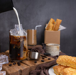 Pour milk to make iced latte coffee, summer coffee in transparent glass, roasted coffee beans, and Crispy Pie on wood. black background studio photo, Copy space for your text