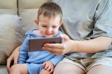 Happy toddler boy sits with his mother and looks into the phone. Mom and child with a smartphone in their hands on the sofa