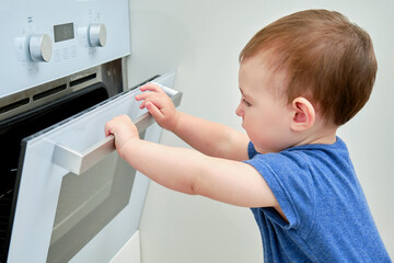 Toddler baby climbs into a hot electric oven. Child boy opens oven door in home kitchen. Kid age...