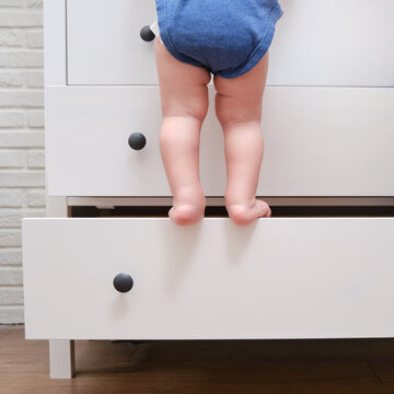 Toddler Baby Climbed Up On The Open Chest Of Drawers. Child Boy Stood On A Tall Drawer Of A White Cabinet. Kid Age One Year