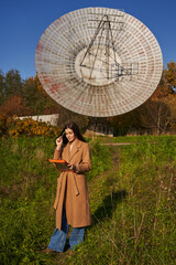 a young woman in a light coat stands near a huge astronomical radar and looks thoughtfully at...