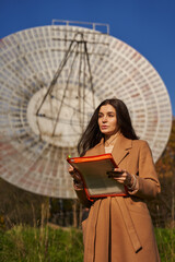 a young woman in a light coat stands near a huge astronomical radar and holds documents in her...