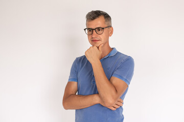 Portrait of thoughtful mature man wearing eyeglasses. Caucasian man wearing blue T-shirt standing with hand on chin over white background. Concentration and uncertainty concept
