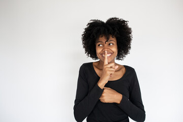 Portrait of cheerful African American woman with finger on lips. Young woman wearing black dress looking away and making silence gesture. Silence and secrecy concept
