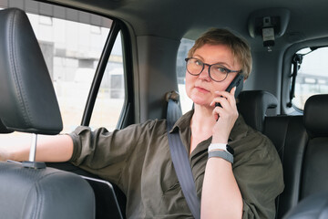 Business woman in glasses is talking on the phone while sitting in the back seat of a car. Business woman is negotiating while riding in a car. Serious middle aged woman with glasses