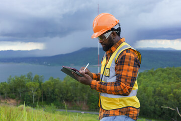 Black Engineer wearing safety jacket and hardhat holding tablet while working at outdoor, Smart working lifestyle.