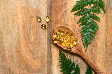 Soft gel omega 3 capsules in the wooden spoon on wooden background with fresh green fern leaves top view, copy space for your text. Natural medicine concept.