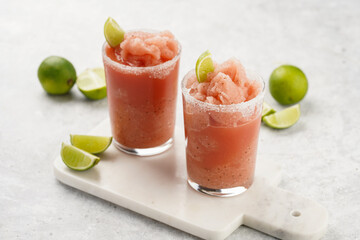 Two glasses with tall long watermelon drink with crushed ice, sugar rims, lime slices on marble board on grey background