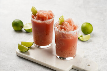 Two glasses with tall long watermelon drink with crushed ice, sugar rims, lime slices on marble board on grey background