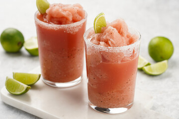 Two glasses with tall long watermelon drink with crushed ice, sugar rims, lime slices on marble board on grey background