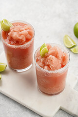 Two glasses with tall long watermelon drink with crushed ice, sugar rims, lime slices on marble board on grey background