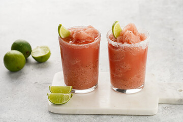 Two glasses with tall long watermelon drink with crushed ice, sugar rims, lime slices on marble board on grey background