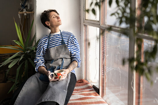Tired female artisan with cup of tea in hands on windowsill, resting in ceramics studio, dreaming to open her own pottery studio. Peaceful female ceramist taking break during workday in workshop - Powered by Adobe