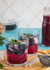 Soft drink with blackberry syrup, sparkling water, whole berries and ice cubes in glasses on a round wooden board on a turquoise concrete background.