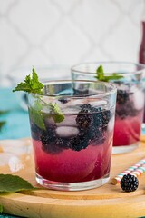 Soft drink with blackberry syrup, sparkling water, whole berries and ice cubes in glasses on a round wooden board on a turquoise concrete background.