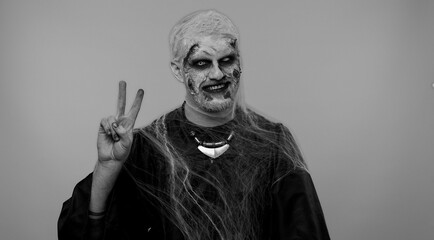 Frightening man with Halloween zombie bloody wounded makeup showing victory sign, hoping for success and win, doing peace gesture, smiling with kind optimistic expression on studio gray