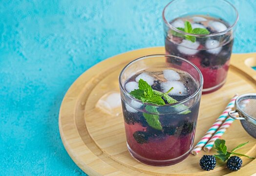 Soft drink with blackberry syrup, sparkling water, whole berries and ice cubes in glasses on a round wooden board on a turquoise concrete background.