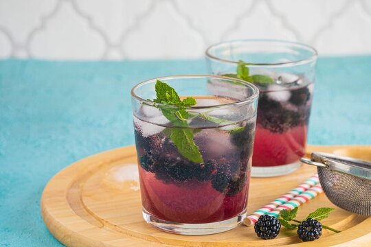 Soft drink with blackberry syrup, sparkling water, whole berries and ice cubes in glasses on a round wooden board on a turquoise concrete background.