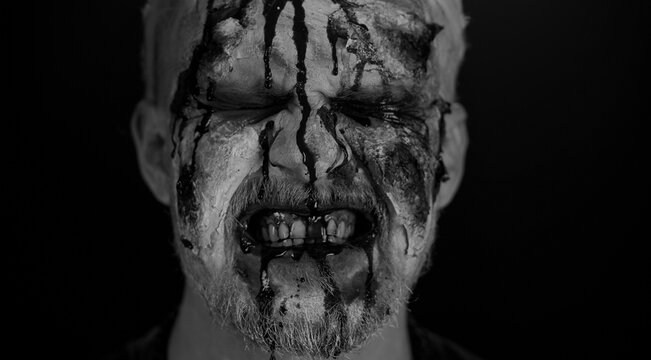 Close-up of sinister man with horrible scary Halloween zombie makeup making faces, looking ominous at camera, trying to scare, blood flows and drips on face. Dead guy with wounded bloody scars face