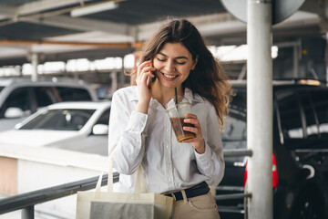 Business young woman with coffee talking cheerfully on the phone in the parking lot.