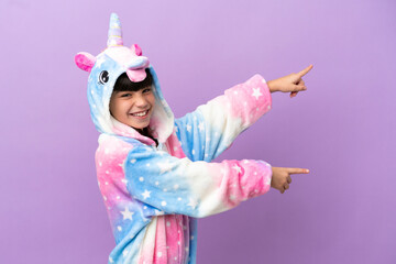 Little kid wearing a unicorn pajama isolated on purple background pointing finger to the side and presenting a product