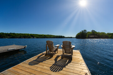 Cottage life - Sunrise on two empty Adirondack chairs sitting on a dock on a lake in Muskoka, Ontario Canada. The sun light create long shadows on the wooden pier. - obrazy, fototapety, plakaty