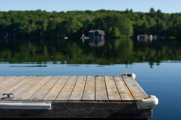 Empty portion of a wooden dock facing a lake in Muskoka Ontario, Canada. Across the the calm waters some cottages are visible nestled between green trees. 