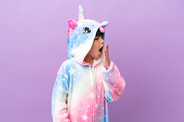 Little kid wearing a unicorn pajama isolated on purple background yawning and covering wide open...
