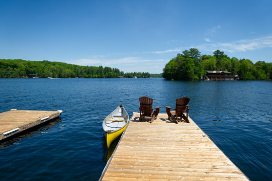 Two Adirondack chairs on a wooden dock facing the blue water of a lake in Muskoka, Ontario Canada. A yellow canoe is tied to the pier.