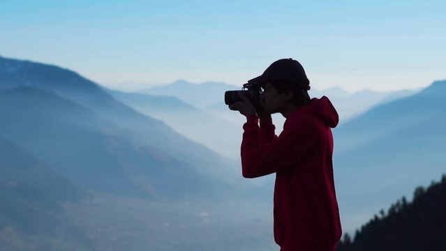 Silhouette shot of an Indian male photographer looking at the viewfinder of the camera in front of the foggy mountains at Manali in Himachal Pradesh, India. Man takes photographs of Himalayas.