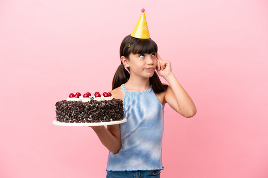 Little caucasian kid holding birthday cake isolated in pink background having doubts and thinking