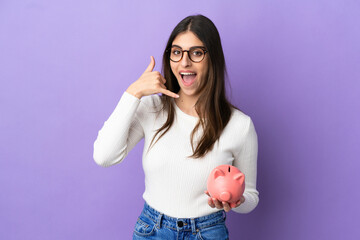 Young caucasian woman holding a piggybank isolated on purple background making phone gesture. Call me back sign