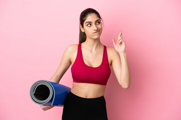 Young sport caucasian woman going to yoga classes while holding a mat with fingers crossing and wishing the best